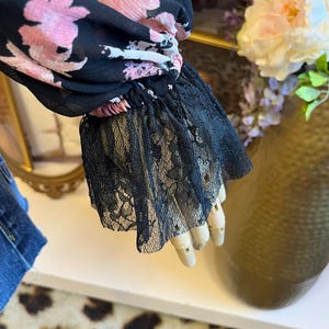 May include: Black floral print blouse with pink flowers and black lace cuffs. The sheer fabric drapes elegantly, showcasing the delicate floral pattern. The lace cuffs add a touch of sophistication to the design.