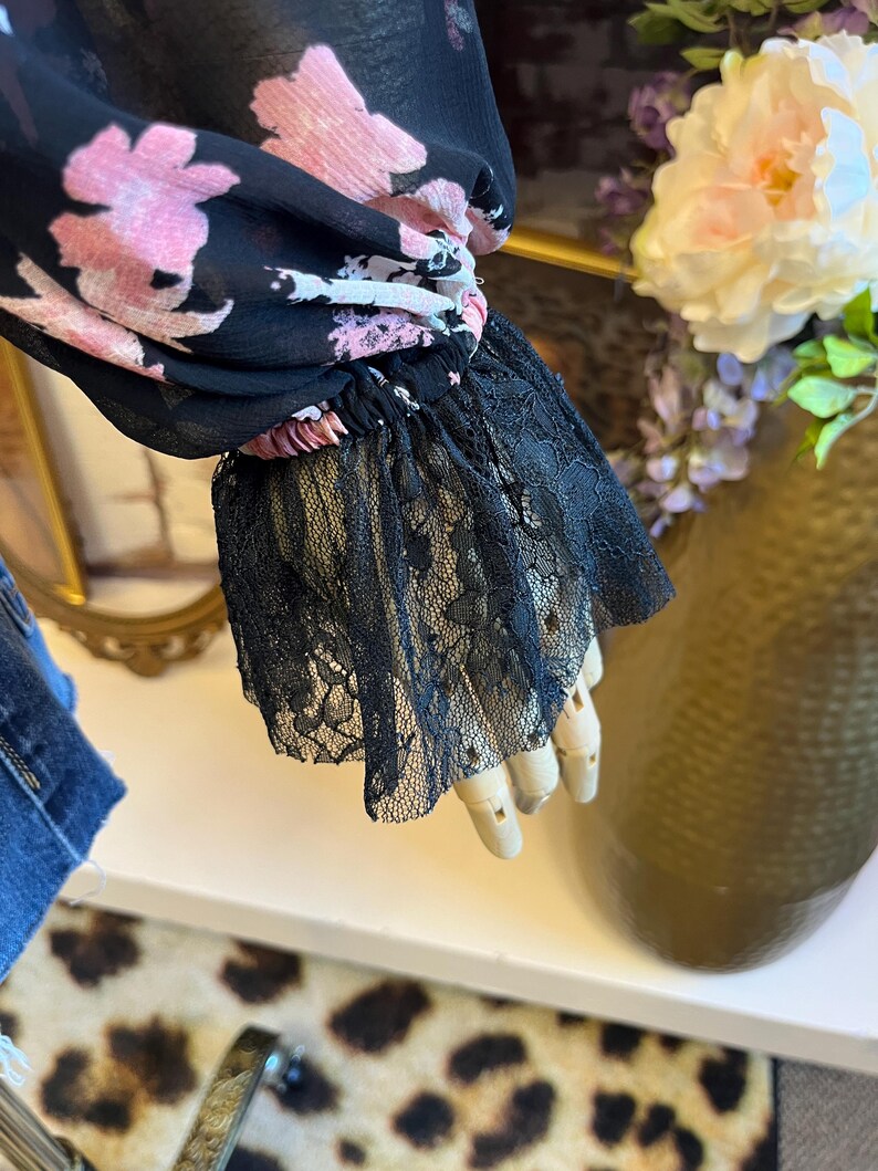 May include: Black floral print blouse with pink flowers and black lace cuffs. The sheer fabric drapes elegantly, showcasing the delicate floral pattern. The lace cuffs add a touch of sophistication to the design.