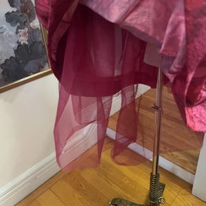 May include: A layered, magenta-colored garment, possibly a dress or skirt, hangs on a metal dress form. The garment features a sheer, flowing overlay, revealing a structured underlayer. The dress form has an ornate, decorative base.