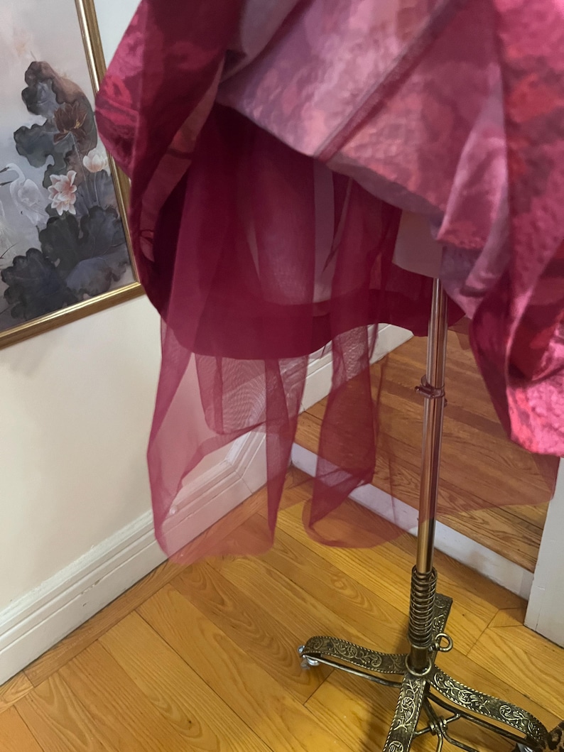 May include: A layered, magenta-colored garment, possibly a dress or skirt, hangs on a metal dress form. The garment features a sheer, flowing overlay, revealing a structured underlayer. The dress form has an ornate, decorative base.