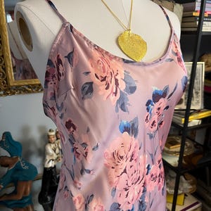May include: A lilac-colored slip dress with a floral pattern of pink and blue roses. The dress has thin straps and is accessorized with a gold leaf pendant necklace. The dress is displayed on a mannequin.