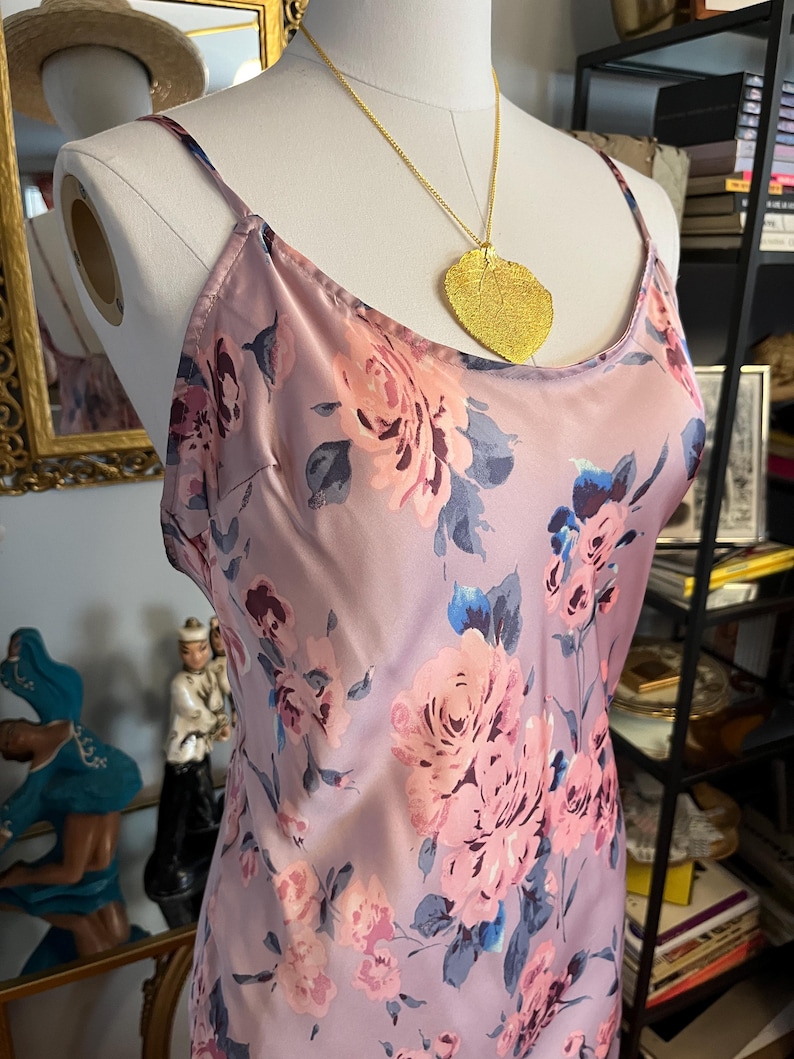 May include: A lilac-colored slip dress with a floral pattern of pink and blue roses. The dress has thin straps and is accessorized with a gold leaf pendant necklace. The dress is displayed on a mannequin.