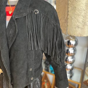 May include: Black suede jacket with fringe detail on the sleeves. The jacket has a notched collar, button closures, and a cropped silhouette. The sleeves are long with a ribbed knit cuff. A silver-tone metal accent is on the shoulder.