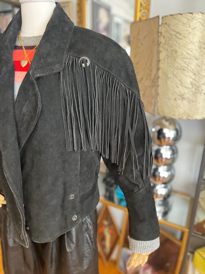 May include: Black suede jacket with fringe detail on the sleeves. The jacket has a notched collar, button closures, and a cropped silhouette. The sleeves are long with a ribbed knit cuff. A silver-tone metal accent is on the shoulder.