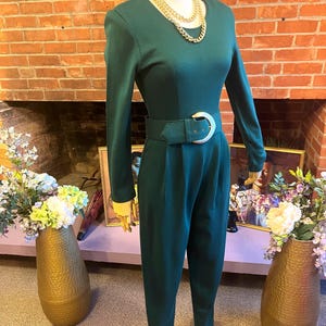 May include: A dark green jumpsuit with long sleeves and a wide belt featuring a large, silver-colored buckle. The jumpsuit has a round neckline and is accessorized with a gold chain necklace. The pants have a stirrup style at the bottom.
