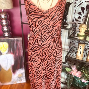 Puede incluir: Un vestido lencero color melocot&oacute;n con un estampado de rayas de cebra negras. El vestido tiene un cuello de cisne y tirantes finos. Un collar dorado en capas est&aacute; drapeado sobre el vestido. El vestido se exhibe en un maniqu&iacute;.