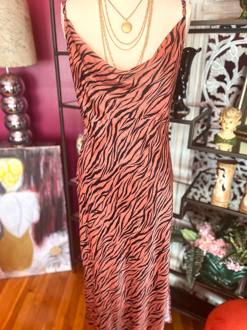 Puede incluir: Un vestido lencero color melocot&oacute;n con un estampado de rayas de cebra negras. El vestido tiene un cuello de cisne y tirantes finos. Un collar dorado en capas est&aacute; drapeado sobre el vestido. El vestido se exhibe en un maniqu&iacute;.