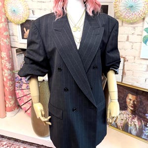 May include: A charcoal gray double-breasted blazer featuring thin vertical pinstripes, wide lapels, and four black buttons. The garment has long sleeves with folded cuffs and two front flap pockets, displayed on a mannequin with layered gold necklaces.