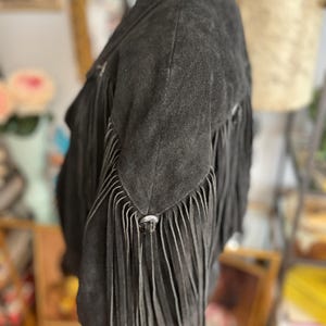 May include: A black suede jacket with long fringe detailing on the sleeves and back. The jacket has a classic collar and silver-toned metal hardware. The fringe adds a Western-inspired flair to the garment.