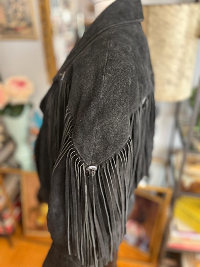 May include: A black suede jacket with long fringe detailing on the sleeves and back. The jacket has a classic collar and silver-toned metal hardware. The fringe adds a Western-inspired flair to the garment.