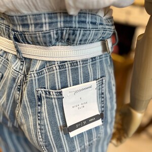 Pode incluir: Jeans slim de cintura alta em azul claro com riscas brancas verticais. Os jeans t&ecirc;m um cinto branco e uma etiqueta que diz "HIGH-RISE SLIM" e "SIZE 31 ANKLE".