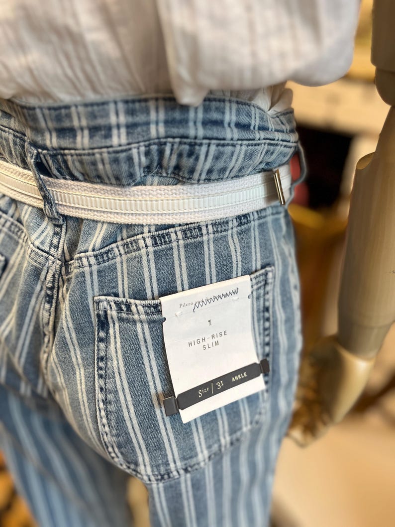 Pode incluir: Jeans slim de cintura alta em azul claro com riscas brancas verticais. Os jeans t&ecirc;m um cinto branco e uma etiqueta que diz "HIGH-RISE SLIM" e "SIZE 31 ANKLE".