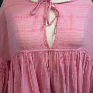 May include: A light pink, loose-fitting top with a keyhole neckline and tie closure. The top features a gathered, tiered design and wide sleeves. The fabric has a subtle striped texture.