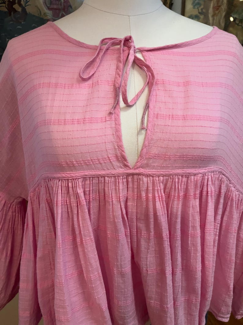 May include: A light pink, loose-fitting top with a keyhole neckline and tie closure. The top features a gathered, tiered design and wide sleeves. The fabric has a subtle striped texture.