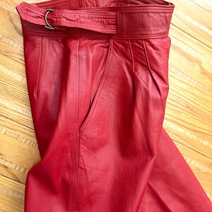 May include: Red leather pants with a high waist and a belt with a silver-colored buckle. The pants have a classic design with a pocket on the side. The leather has a smooth texture and a vibrant red color.