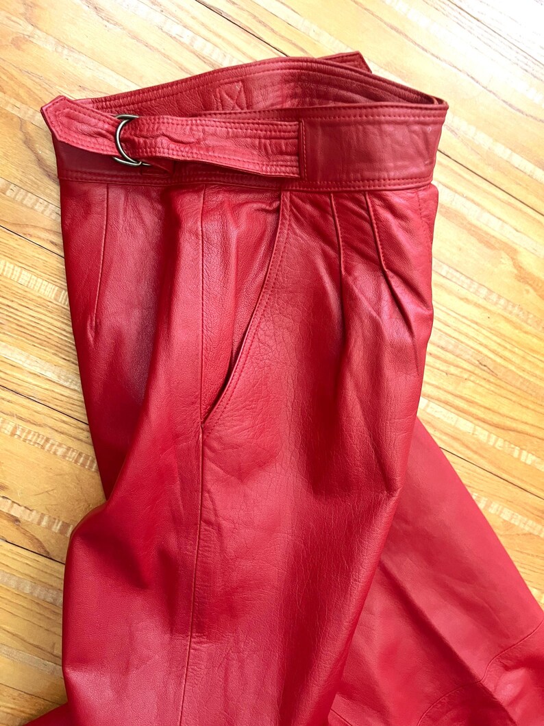 May include: Red leather pants with a high waist and a belt with a silver-colored buckle. The pants have a classic design with a pocket on the side. The leather has a smooth texture and a vibrant red color.