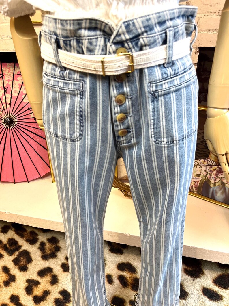 Pode incluir: Cal&ccedil;as de ganga azul claro com riscas brancas verticais. As cal&ccedil;as apresentam cintura alta, braguilha com bot&otilde;es e bolsos frontais. Um cinto branco com fivela dourada aperta a cintura. As cal&ccedil;as s&atilde;o exibidas num manequim.