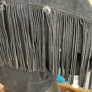 May include: Close-up of a black suede jacket with long fringe detailing. The jacket features silver-toned button closures and a soft, textured appearance. The fringe hangs vertically, adding a Western-inspired aesthetic.