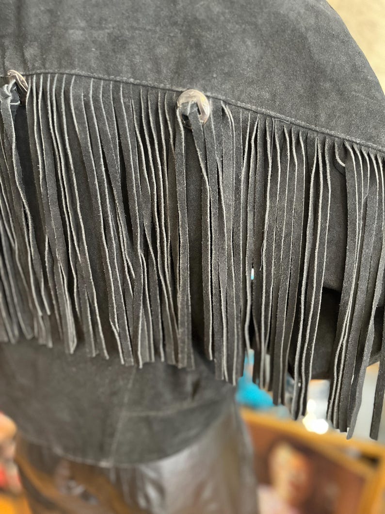 May include: Close-up of a black suede jacket with long fringe detailing. The jacket features silver-toned button closures and a soft, textured appearance. The fringe hangs vertically, adding a Western-inspired aesthetic.