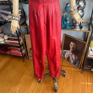 May include: Bright red leather pants with a high waist and pleated front. The pants are displayed on a mannequin with a black top and gold-toned jewelry. The pants have a classic, tailored fit.