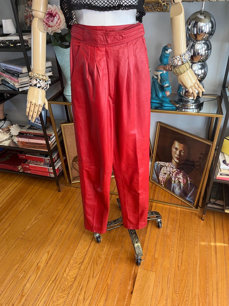 May include: Bright red leather pants with a high waist and pleated front. The pants are displayed on a mannequin with a black top and gold-toned jewelry. The pants have a classic, tailored fit.