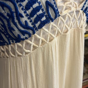 Pu&ograve; includere: Primo piano di un vestito color crema con dettagli ricamati blu sulle maniche. L'abito presenta uno scollo a V e un tessuto strutturato. Il design include elementi traforati all'uncinetto.