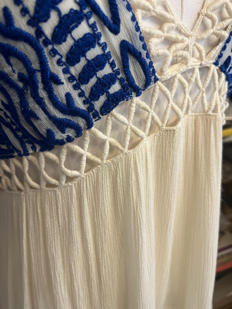 Pu&ograve; includere: Primo piano di un vestito color crema con dettagli ricamati blu sulle maniche. L'abito presenta uno scollo a V e un tessuto strutturato. Il design include elementi traforati all'uncinetto.