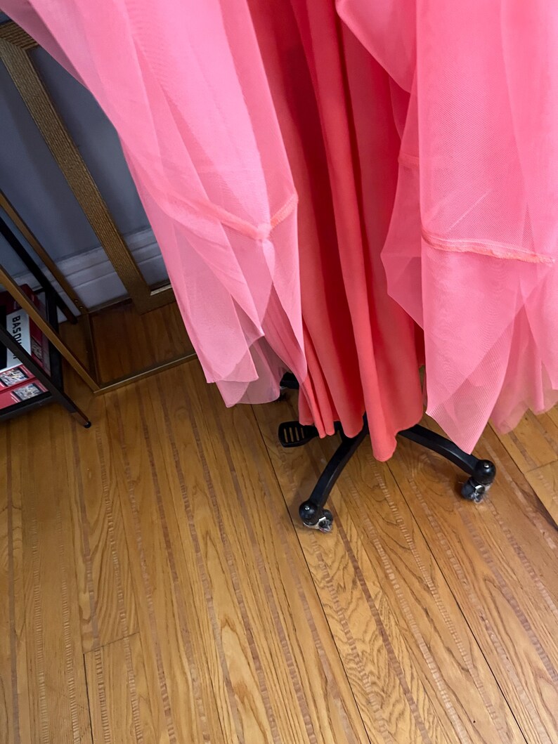 Puede incluir: Un vestido fluido de color coral con capas de tul rosa transparente. El vestido se exhibe sobre un maniqu&iacute; negro con ruedas, sobre un fondo de suelo de madera. El tul est&aacute; recogido y cosido en los bordes.