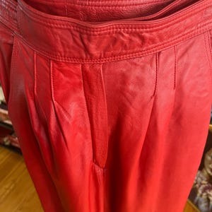 May include: Close-up of bright red leather pants. The pants have a high waist, pleats, and a tailored fit. The leather appears smooth and has a slight sheen. The pants are displayed on a mannequin.