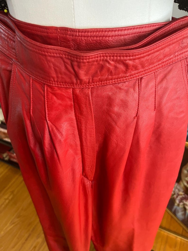 May include: Close-up of bright red leather pants. The pants have a high waist, pleats, and a tailored fit. The leather appears smooth and has a slight sheen. The pants are displayed on a mannequin.