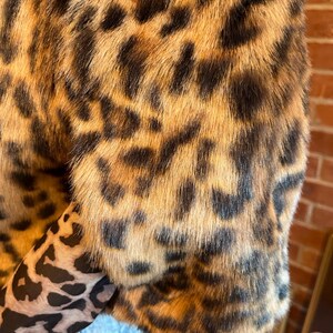 K&ouml;nnte beinhalten: Eine kurz geschnittene Jacke aus Kunstfell mit einem braun-beigen Leopardenmuster. Das Kleidungsst&uuml;ck hat kurze &Auml;rmel und wird &uuml;ber einem lang&auml;rmeligen Oberteil mit einem kleineren schwarz-beigen Leopardenmuster sowie einer hellblauen Jeans getragen.