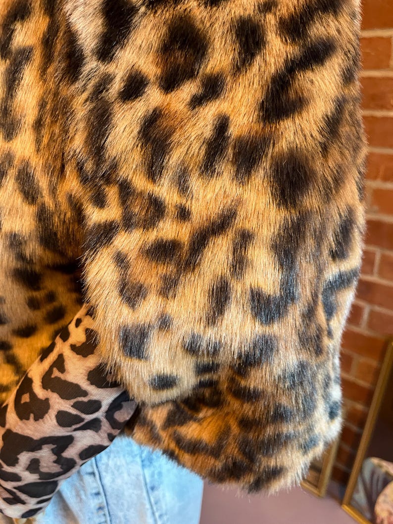 K&ouml;nnte beinhalten: Eine kurz geschnittene Jacke aus Kunstfell mit einem braun-beigen Leopardenmuster. Das Kleidungsst&uuml;ck hat kurze &Auml;rmel und wird &uuml;ber einem lang&auml;rmeligen Oberteil mit einem kleineren schwarz-beigen Leopardenmuster sowie einer hellblauen Jeans getragen.