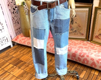 May include: Light blue denim jeans featuring a patchwork design with rectangular panels in varying shades of blue and white. The jeans have a brown leather belt with a silver-tone metal buckle and are cuffed at the bottom of the pant legs.