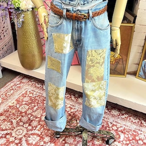 May include: Light blue denim jeans featuring a brown braided leather belt and multiple rectangular patches of gold floral brocade fabric sewn onto the front of the legs. The jeans have a straight-leg fit with cuffed hems at the bottom.