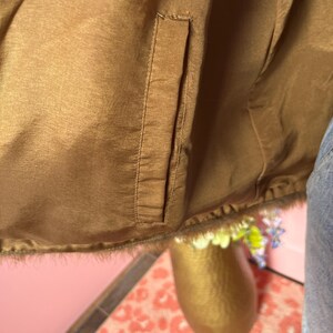 May include: Close-up of a brown satin jacket with a pocket. The jacket has a fur trim at the bottom. The background includes a gold vase with flowers and a pink wall.