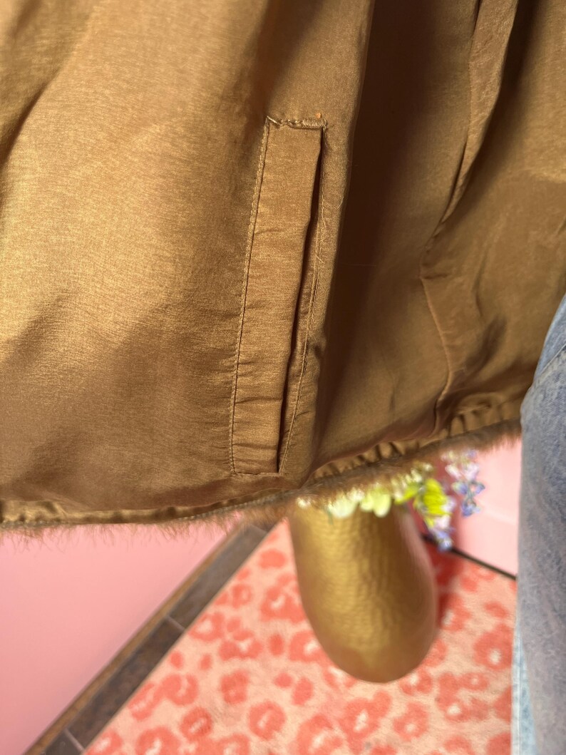 May include: Close-up of a brown satin jacket with a pocket. The jacket has a fur trim at the bottom. The background includes a gold vase with flowers and a pink wall.
