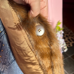 May include: Close-up of a brown satin fabric with a fur trim and a large, round, silver button. The fur is a mix of brown and black. The satin fabric is a warm, golden brown color.