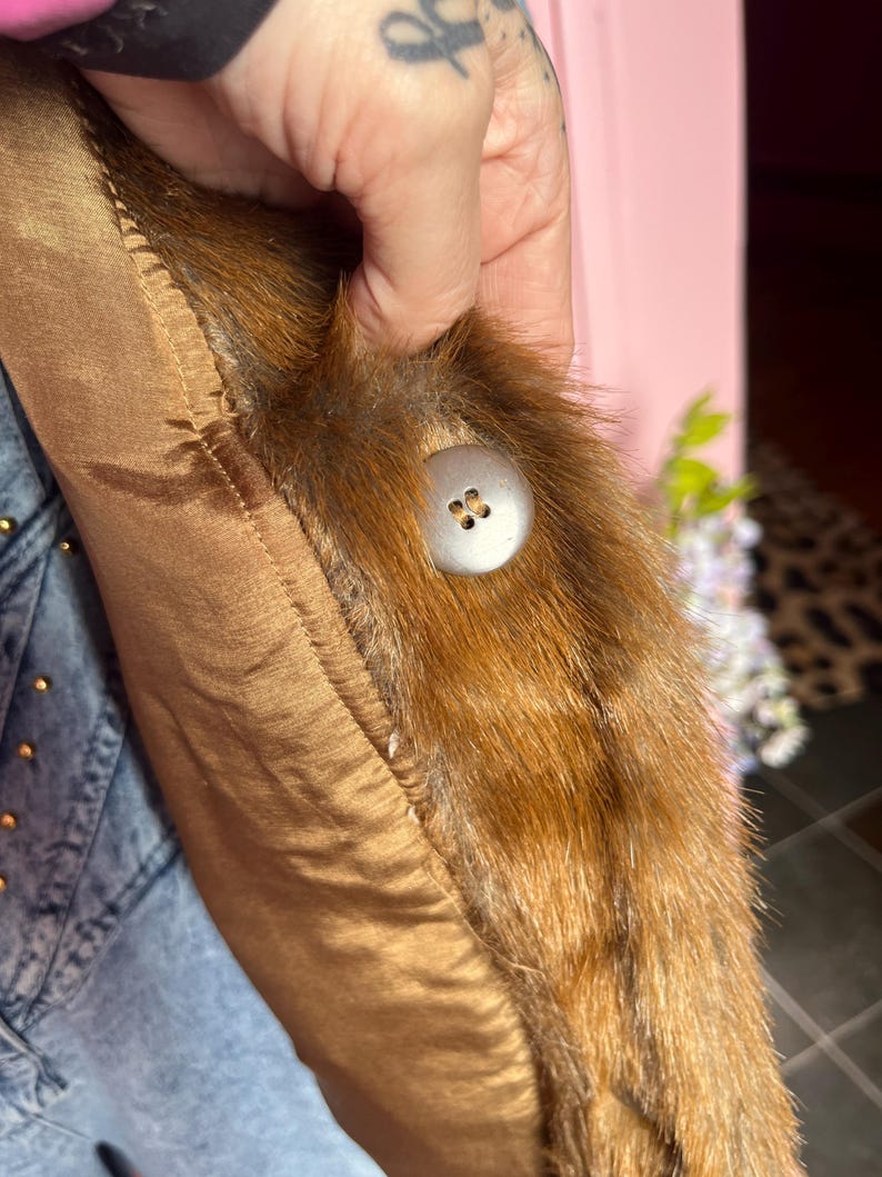 May include: Close-up of a brown satin fabric with a fur trim and a large, round, silver button. The fur is a mix of brown and black. The satin fabric is a warm, golden brown color.