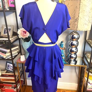 May include: A vibrant blue dress with a V-neckline and a button closure. The dress features a cut-out detail at the waist and a tiered, ruffled skirt. A gold-colored belt cinches the waist. The dress is displayed on a mannequin.