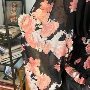 May include: Black blouse with sheer sleeves featuring a large pink floral pattern. The top has a gathered waist and a black lace trim at the cuffs. The floral design includes shades of pink and white.