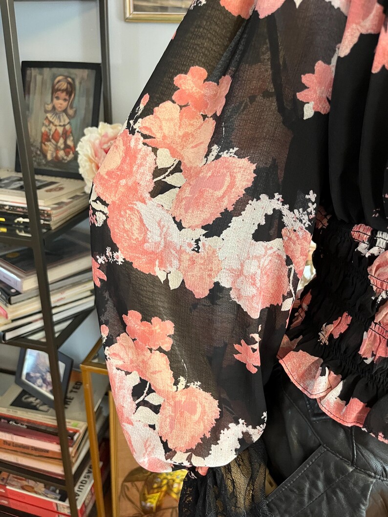 May include: Black blouse with sheer sleeves featuring a large pink floral pattern. The top has a gathered waist and a black lace trim at the cuffs. The floral design includes shades of pink and white.