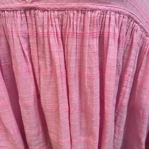 May include: A light pink blouse with a V-neck and textured fabric. The top features a gathered design below the neckline, creating a flowing silhouette. The fabric has a subtle grid pattern and horizontal stripes, adding visual interest to the garment.