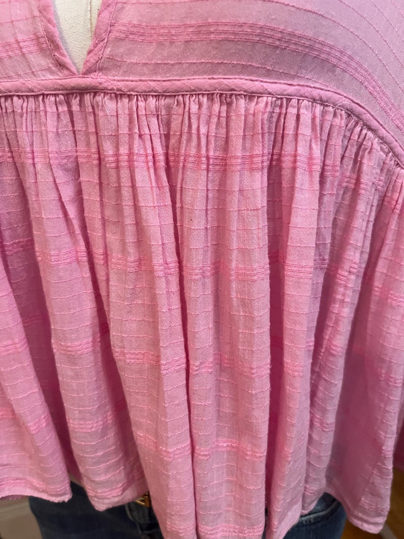 May include: A light pink blouse with a V-neck and textured fabric. The top features a gathered design below the neckline, creating a flowing silhouette. The fabric has a subtle grid pattern and horizontal stripes, adding visual interest to the garment.
