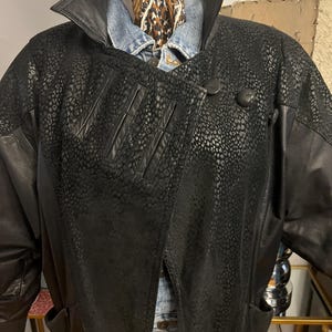 May include: Black leather jacket with a unique spotted pattern on the front and sleeves. The jacket has a large collar, button closure, and pockets. A denim shirt and patterned scarf are visible underneath the jacket.