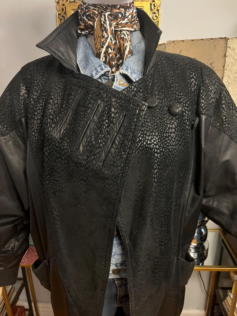 May include: Black leather jacket with a unique spotted pattern on the front and sleeves. The jacket has a large collar, button closure, and pockets. A denim shirt and patterned scarf are visible underneath the jacket.