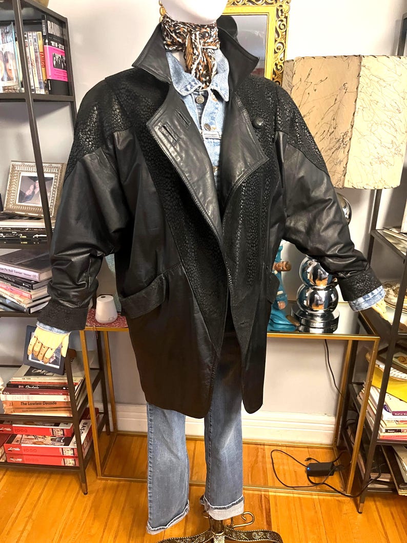 May include: A black leather jacket with textured panels on the shoulders and upper sleeves. The jacket has a wide collar, a button closure, and two front pockets. The jacket is paired with a denim shirt and blue jeans.