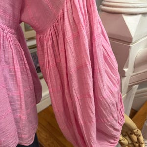 May include: A pink blouse with long, voluminous sleeves. The fabric has a textured, striped pattern. The sleeves are gathered at the wrist, creating a puffy effect. The blouse is a solid, light pink color.