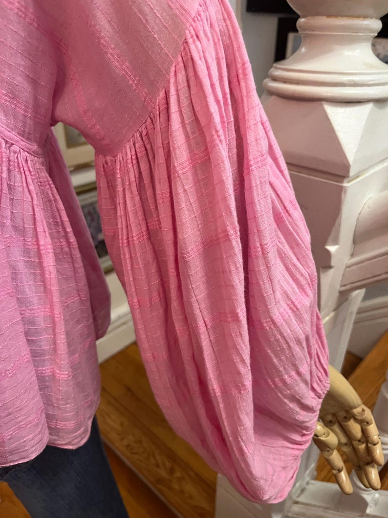 May include: A pink blouse with long, voluminous sleeves. The fabric has a textured, striped pattern. The sleeves are gathered at the wrist, creating a puffy effect. The blouse is a solid, light pink color.