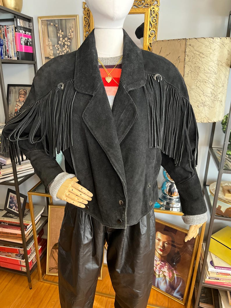 May include: Black suede jacket with fringe detail on the sleeves. The jacket has a classic collar, button closure, and is paired with black leather pants. The mannequin is wearing a striped sweater and a gold necklace.