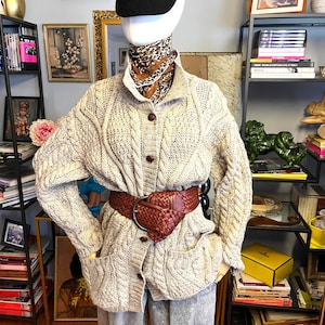 May include: A mannequin wearing a beige cable-knit cardigan with a brown leather belt and gray pants. The mannequin also has a black corduroy cap and a patterned scarf. The cardigan has two front pockets and wooden buttons.
